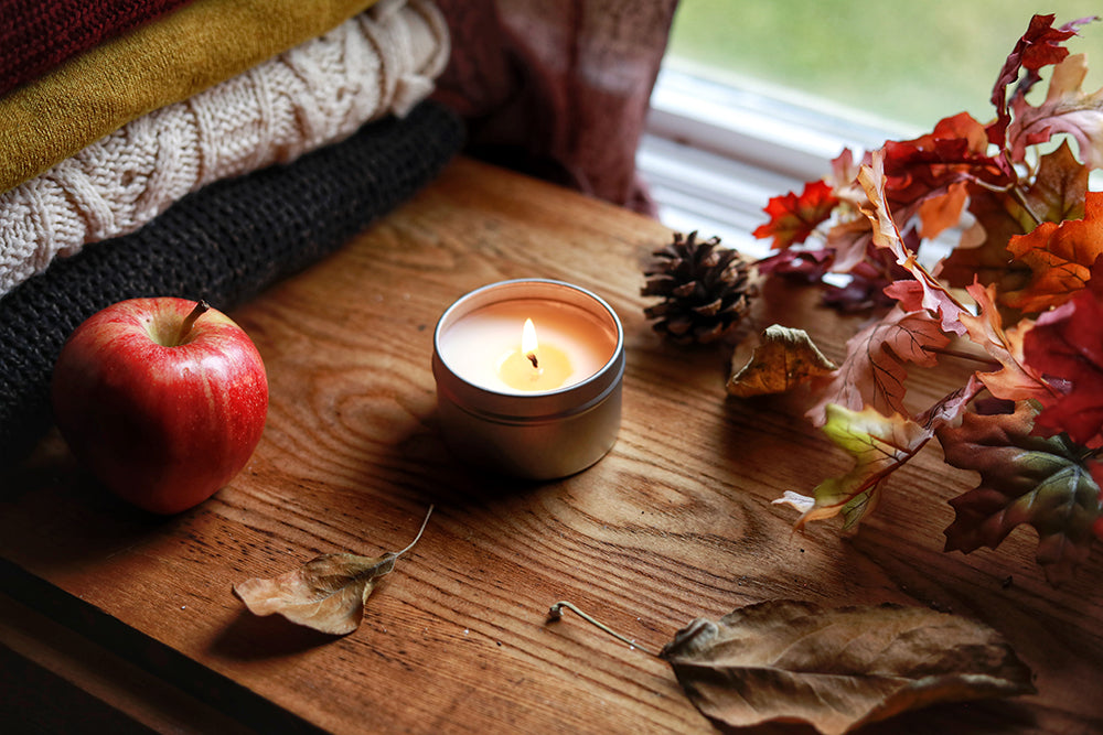A Skinny Dip Candle sits on a wooden table near a window, dried leaves, cozy sweaters and an apple.