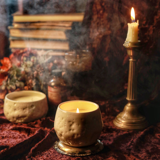 A candle in an off-white ceramic jar made to look like the surface of the moon sits in the center of a red-velvet-clad table. A lit taper candle, a stack of old books and dried flowers are slightly blurry behind it.