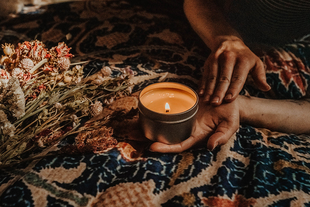 A hand holds a lit Skinny Dip Candle while the other hand rubs the wax into their skin. The background is a colorful design and dried flower bouquet.