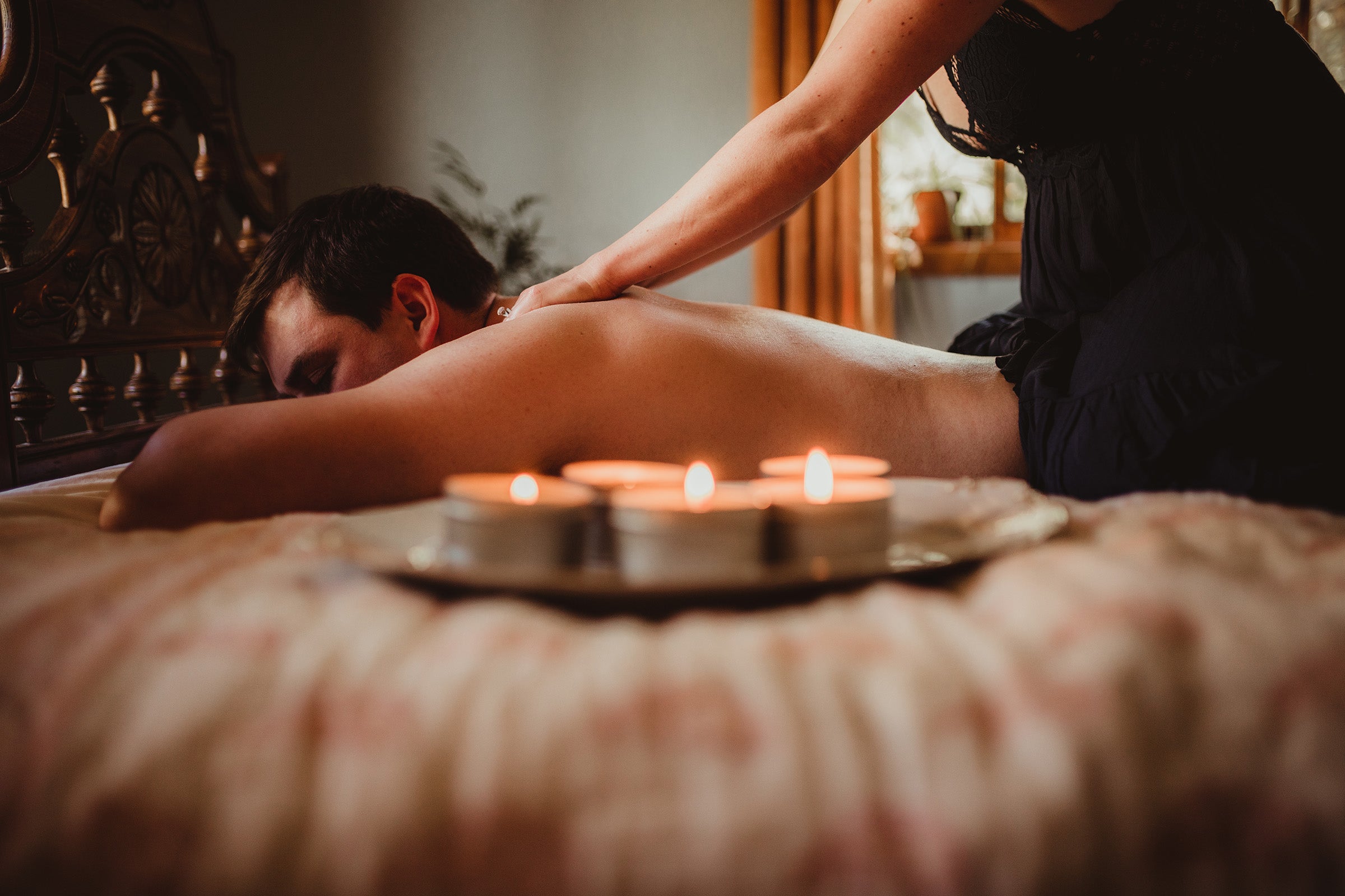 A man lays facedown on a bed as a woman massages him. In the foreground is a plate full of lit Skinny Dip Candles.