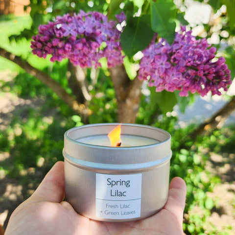 A hand holds out a lit Skinny Dip Candle with a lilac bush in the background. The label reads, "Spring Lilac - fresh lilac & green leaves."