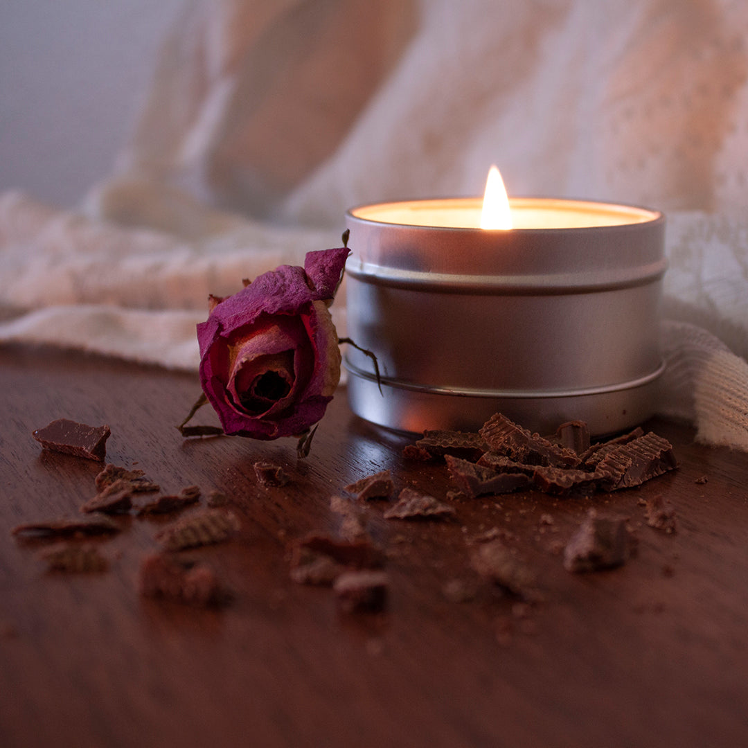 A lit Skinny Dip Candle sits on a mahogany table with white lace in the background. Chocolate shavings are scattered around the candle and a dried pink rose is next to it.