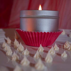 A lit Skinny Dip Candle sits in dark pink cupcake holders. Buttercream frosting dollops are scattered on a plate surrounding it.