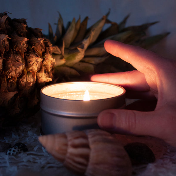 A lit Skinny Dip Candle sits in a surrounding of a pineapple, shells and shredded coconut. A hand grips the candle as if to pick it up.