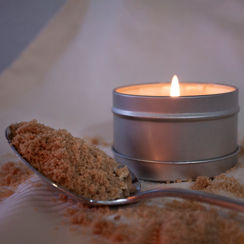 A silver spoon of brown sugar rests in front of a lit Skinny Dip Candle. Sugar litters the pillowcase surface.
