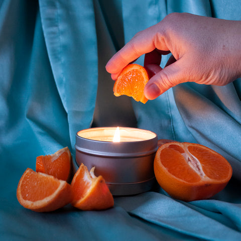 A lit Skinny Dip Candle sits on a soft blue satin robe. Surrounding it are tangerine wedges. A hand squeezes a slice above the candle.