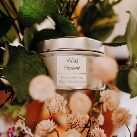 A Skinny Dip Candle in a tin jar sits in a bouquet of pink flowers and green foliage. The label reads, "Wild Flower: Lilac, Sweet Pea, Clay and Sandalwood."